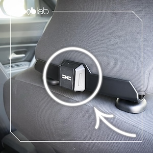 May include: A black plastic car headrest mount featuring a central rectangular housing with a silver side panel and a white DC logo on the front. The device is securely attached to the base of a dark gray fabric car headrest, providing a stable support.