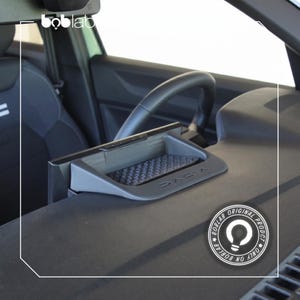 Puede incluir: Una bandeja gris para el salpicadero del coche con la marca "DACIA" visible. La bandeja tiene una superficie texturizada y está situada cerca del volante. La imagen también muestra un logotipo "BOBLAB" y texto que indica que es un producto original.