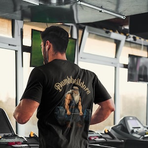 May include: A person wearing a black t-shirt with a gold graphic of a muscular man lifting weights. The text "Dumbbelldora" is printed on the shirt. The person is running on a treadmill in a gym.