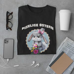 Può includere: T-shirt nera con la scritta "PIXELIGE OSTERN" sopra un coniglietto pixelato con un fiocco rosa e un uovo di Pasqua. Il coniglietto è circondato da fiori e un bordo blu smerlato. Altri oggetti includono un telefono, occhiali e una tazza di caffè.