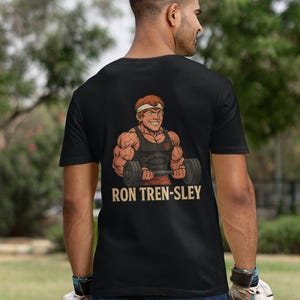 Pode incluir: Camiseta preta com uma ilustração de desenho animado de um homem musculoso levantando pesos. O texto "RON TREN-SLEY" é impresso abaixo da ilustração.