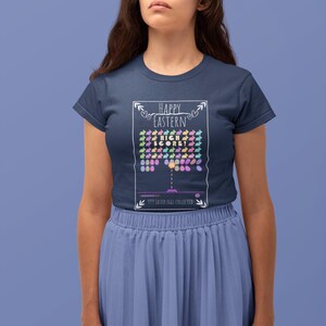 May include: A navy blue t-shirt with a white graphic of a video game screen. The graphic says "Happy Easter" and "High Score!" with a depiction of a game where the player collects Easter eggs. The text at the bottom of the graphic says "9999 Easter Eggs Collected."
