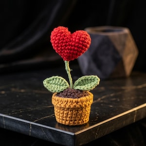 Könnte beinhalten: Eine kleine Häkelpflanze mit einer leuchtend roten herzförmigen Blüte an einem grünen Stiel mit zwei grünen Blättern. Die Pflanze steht in einem strukturierten braunen Häkeltopf mit dunkelbrauner Erde, ein kompaktes und detailliertes Handarbeitsobjekt.