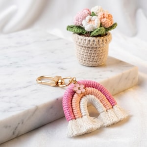 May include: A handmade rainbow keychain with pink, peach, and white colors, featuring a gold clasp and a small pink flower charm. A crocheted flower pot with pink and white flowers sits in the background. The keychain is a charming accessory.