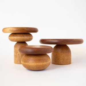 May include: Three wooden decorative stands in varying sizes and shapes. The stands are made of light and dark brown wood. One stand has a stacked design, while the others have a pedestal base with a round top. The background is white.