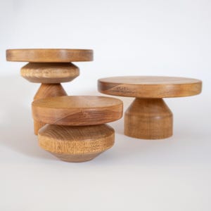 Peut inclure: Quatre présentoirs en bois de différentes hauteurs et formes. Les présentoirs sont en bois brun clair avec une finition naturelle. Ils ont des dessus ronds et des bases de différentes formes, dont empilées et coniques. Le fond est blanc uni.