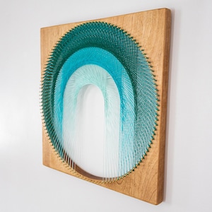 May include: Wooden wall art with a string art design. The artwork features an arch shape made with teal and light blue threads. The threads are arranged around small gold pins, creating a gradient effect. The wooden frame is a light brown color.