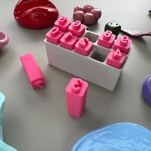 Peut inclure: Un ensemble de tampons numériques roses dans un support blanc, avec d'autres tampons roses et des formes abstraites colorées. Les tampons affichent les chiffres de 0 à 9. D'autres objets incluent un objet rouge brillant en forme de cœur et un cube vert.