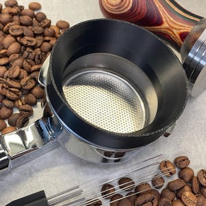 Puede incluir: Un portafiltro cromado para máquina de espresso con un inserto negro, rodeado de granos de café tostado. También se ven un tamper de madera y un distribuidor de agujas para café, lo que sugiere el tema de la preparación del café.