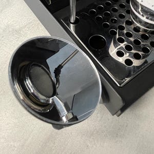 May include: A close-up shot of a chrome espresso machine portafilter with a mesh filter. The machine has a black base and a stainless steel drip tray. A cup is partially visible on the tray. The image is taken from above.