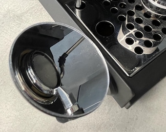 May include: Close-up of a chrome espresso machine portafilter with a mesh filter. The machine has a black base and a stainless steel drip tray. A partially visible cup sits on the tray. The image is taken from a high angle.
