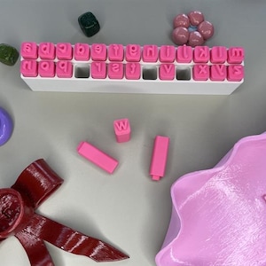 Puede incluir: Una colección de sellos de letras rosas en una bandeja blanca, con sellos rosas adicionales y una forma ondulada rosa. También se incluyen un lazo rojo, piedras decorativas moradas y verdes y un plato morado.