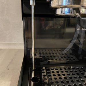 Op de afbeelding: Close-up van een zwarte espressomachine met een roestvrijstalen lekbak en een doorzichtig waterreservoir. Een lange, zilveren metalen staaf loopt naar beneden. De machine staat op een lichtgrijze ondergrond.