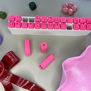 Peut inclure: Un ensemble de tampons lettres roses dans un support blanc, avec d'autres fournitures d'artisanat. Les tampons sont disposés par ordre alphabétique. Autres éléments : un nœud rouge, un cercle violet et des formes roses.