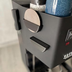 Könnte beinhalten: Eine schwarze Espressomaschine mit einem silbernen Siebträger und einem kleinen roten Licht. Die Maschine hat einen eingebauten Tassenhalter mit einer schwarz-weiß gemusterten Tasse und einem blauen Glas. Ein runder Netzfilter ist an der Seite befestigt.