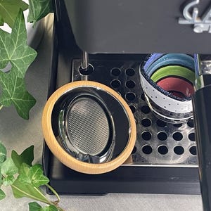 Può includere: Primo piano di una macchina per caffè espresso con un portafiltro e una tazza di ceramica colorata. Il portafiltro ha un manico in legno e un cestello filtro in acciaio inossidabile. La tazza ha uno sfondo bianco con strisce colorate.