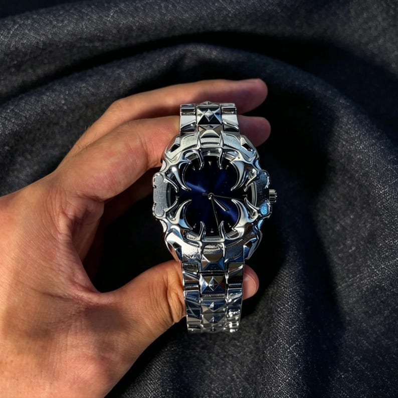 May include: A silver-toned wristwatch with a unique, ornate design. The watch features a dark blue face and a matching silver band. The watch is held in a hand against a dark gray background.