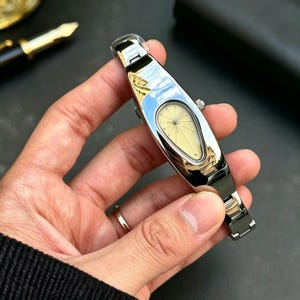 以下が含まれることがあります： ユニークな細長い楕円形の文字盤と淡い黄色の文字盤を備えた、シルバーカラーの腕時計。時計は磨かれた金属製のバンドと小さなリューズを備えています。時計は手で持たれており、その反射面が示されています。
