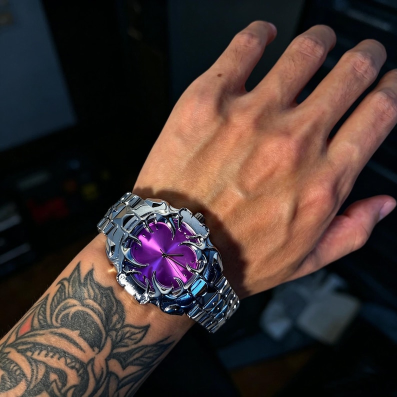 May include: A silver-toned wristwatch with a unique, ornate design. The watch face is a vibrant purple, and the band is made of linked metal segments. The watch is on a wrist with a tattoo.