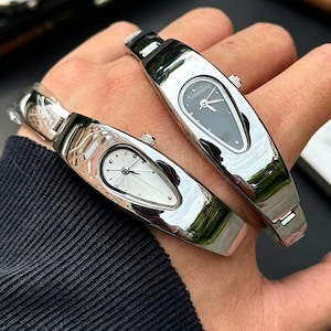 以下が含まれることがあります： ブレスレットデザインの、銀色の楕円形の文字盤の時計が2つ。1つは白い文字盤、もう1つは濃い灰色の文字盤です。時計は人の手に表示され、その洗練されたモダンなデザインを披露しています。時計はミニマリストな美学を持っています。