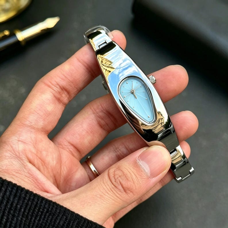以下が含まれることがあります： 水色の涙型の文字盤が付いたシルバーの腕時計。時計はシルバーのバンドとミニマリストのデザインです。文字盤にはサンバースト模様とシルバーの針が付いています。