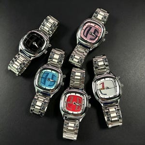 Puede incluir: Cinco relojes de pulsera de acero inoxidable con esferas cuadradas y correas de metal a juego. Cada reloj tiene una esfera de diferente color: negro, rosa, azul, rojo y blanco. Las esferas de los relojes tienen dise&ntilde;os gr&aacute;ficos &uacute;nicos.