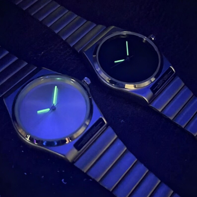 May include: Two minimalist watches with silver bands and dark faces. The watch hands and markers glow green. The watches are set against a dark background, highlighting their sleek design. The watches appear to be made of metal.