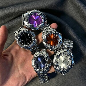 May include: Five silver-toned wristwatches with unique, textured designs. Each watch features a different colored face: purple, black, orange, blue, and silver. The watches are held in a hand, showcasing their intricate details.