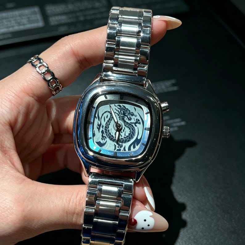 May include: A silver-toned analog wristwatch featuring a square-shaped case with rounded corners and a metallic link bracelet. The watch face displays a detailed black dragon illustration, silver hour markers, and silver hands for timekeeping.