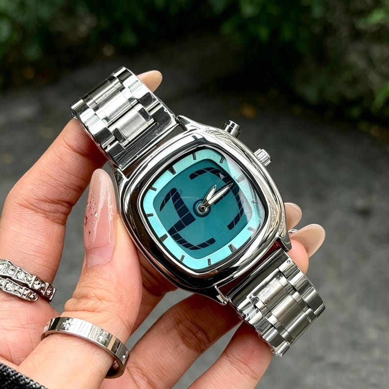 Puede incluir: Un reloj de pulsera plateado con una esfera cuadrada de color turquesa y una correa plateada a juego. La esfera del reloj presenta un dise&ntilde;o negro y agujas plateadas. El reloj se muestra sobre un fondo verde borroso.