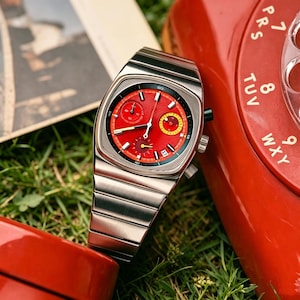 May include: A stainless steel wristwatch with a red face and a silver band. The watch has a square face with multiple dials and a date window. The watch is set against a backdrop of green grass and a red rotary phone.