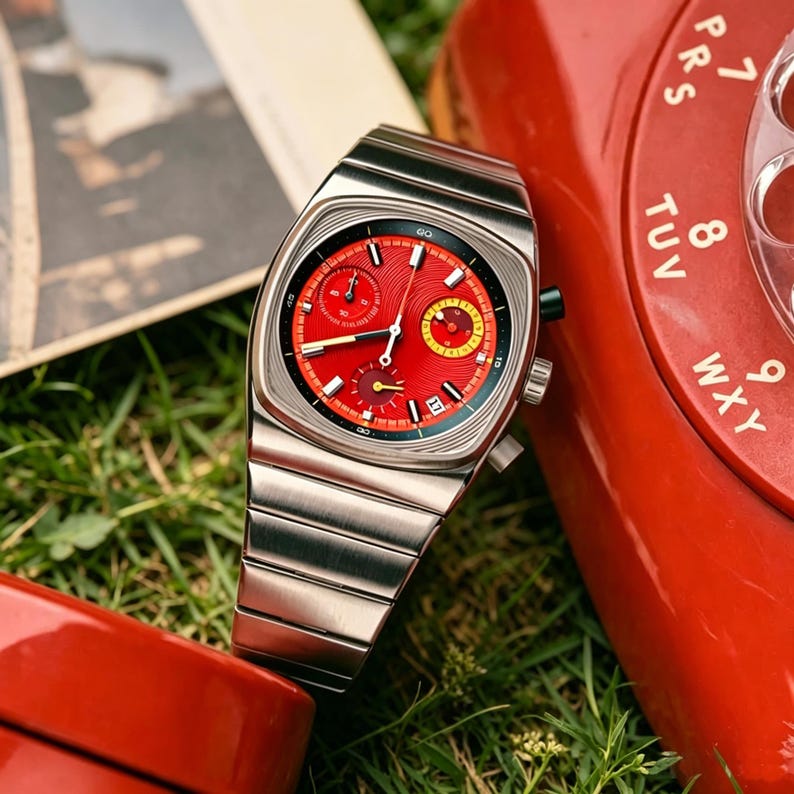 May include: A stainless steel wristwatch with a red face and a silver band. The watch has a square face with multiple dials and a date window. The watch is set against a backdrop of green grass and a red rotary phone.