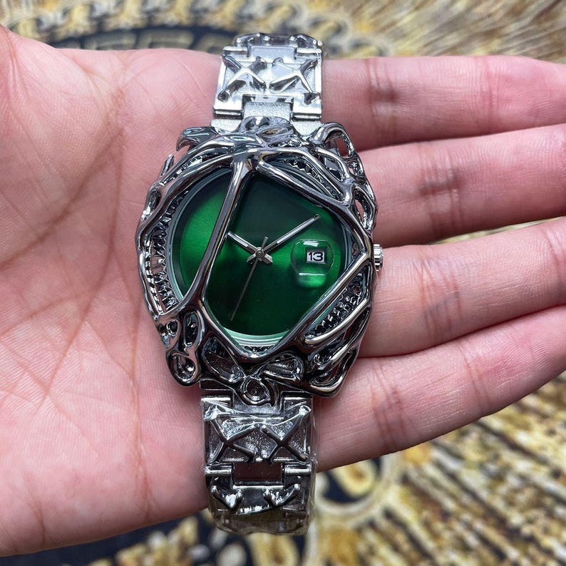 May include: A silver-toned wristwatch with a unique, openwork design around a vibrant green watch face. The watch features a date display at the 3 o'clock position and a matching silver band. The watch is a stylish accessory.
