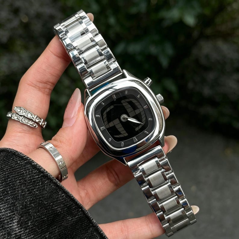 Puede incluir: Un reloj de pulsera plateado con una esfera negra cuadrada y una correa de metal a juego. La esfera del reloj presenta un dise&ntilde;o &uacute;nico. El reloj lo sostiene una persona.
