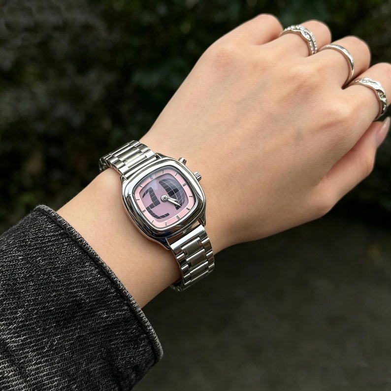 Puede incluir: Un reloj de pulsera plateado con una esfera rosa cuadrada y una correa plateada a juego. La esfera del reloj presenta un dise&ntilde;o estilizado. El reloj se lleva en una mu&ntilde;eca con varios anillos plateados.