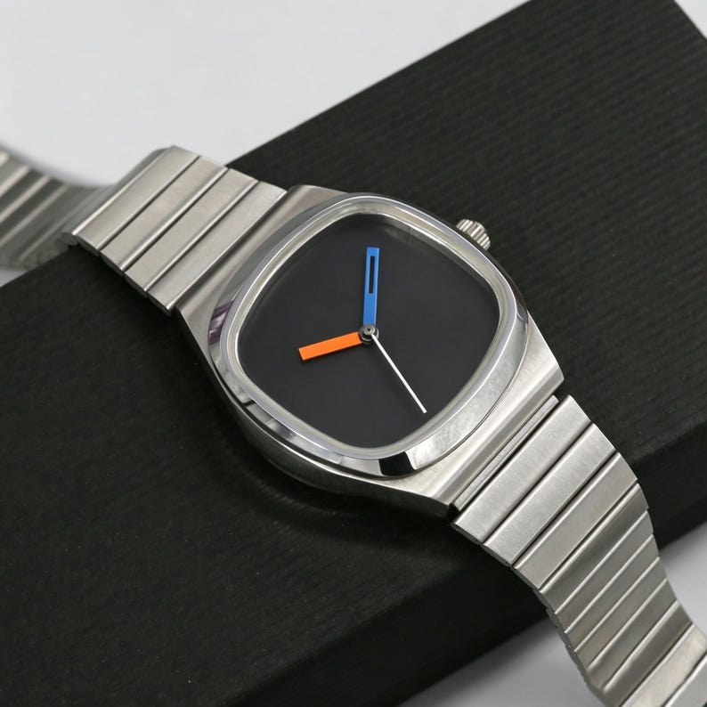 May include: A stainless steel wristwatch with a square black face. The watch features a silver band and orange and blue hands. The watch is displayed on a black surface.