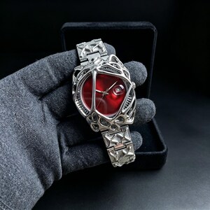 May include: A silver-toned wristwatch with a unique, open-worked design. The watch features a vibrant red face and a date window. The band and case have a geometric pattern. The watch is presented in a black box.