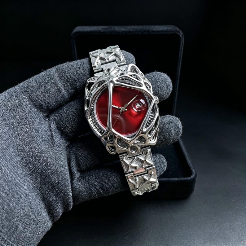 May include: A silver-toned wristwatch with a unique, open-worked design. The watch features a vibrant red face and a date window. The band and case have a geometric pattern. The watch is presented in a black box.