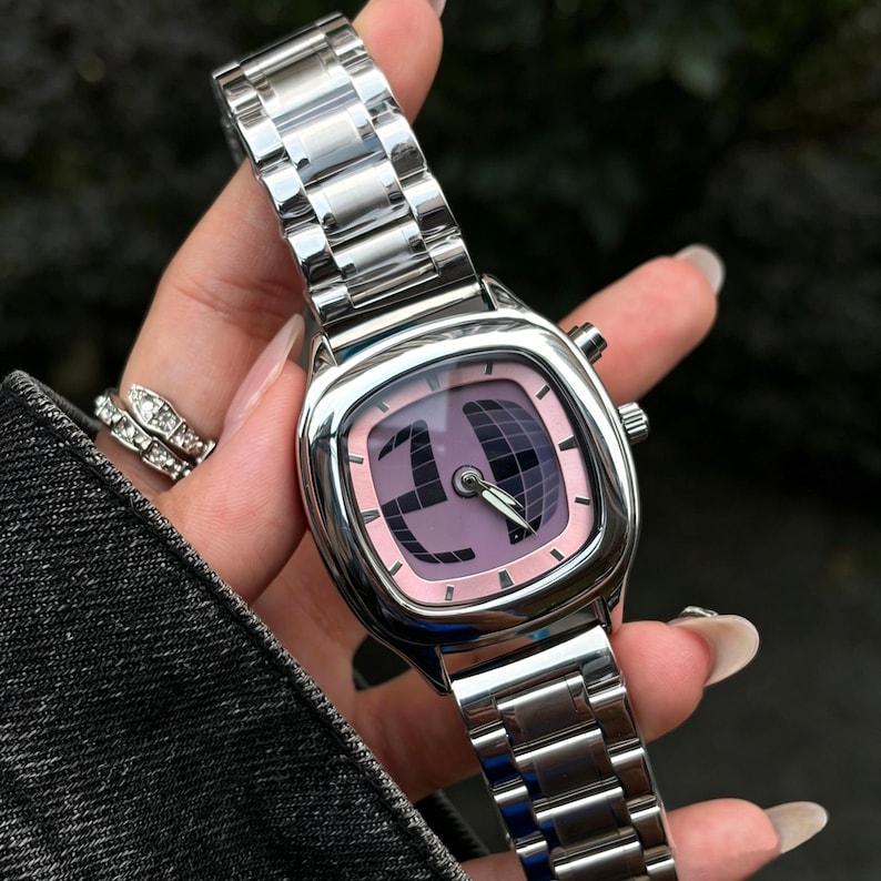 Puede incluir: Un reloj de pulsera plateado con una esfera cuadrada y una correa de metal a juego. La esfera del reloj es de color rosa claro con un dise&ntilde;o gr&aacute;fico oscuro y agujas plateadas. Un accesorio elegante.