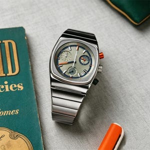 May include: A silver-toned wristwatch with a square face and a silver band. The watch face is gray with blue and orange accents. The watch has two small dials and an orange button on the side. The watch is on a gray surface.