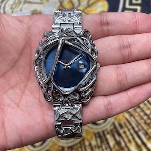 May include: A silver-toned wristwatch with a unique cage-like design around a blue watch face. The watch features a date display and a matching silver band. The watch is held in a hand.