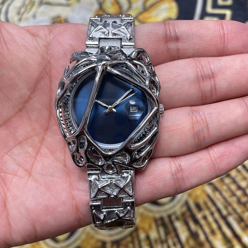 May include: A silver-toned wristwatch with a unique cage-like design around a blue watch face. The watch features a date display and a matching silver band. The watch is held in a hand.