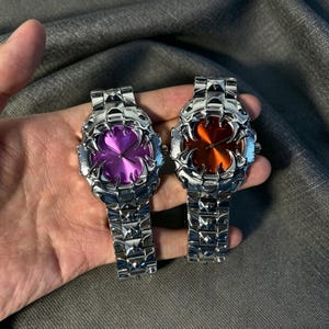 May include: Two silver-toned wristwatches with unique, spiky bezels and matching link bands. One watch has a purple face, the other an orange face. The watches are held against a gray fabric background.