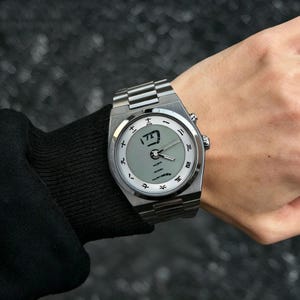 May include: A silver-toned stainless steel wristwatch featuring a light gray circular dial with black Japanese kanji numerals as hour markers. The watch includes a metallic link bracelet, silver hands, and a prominent kanji character at the center.