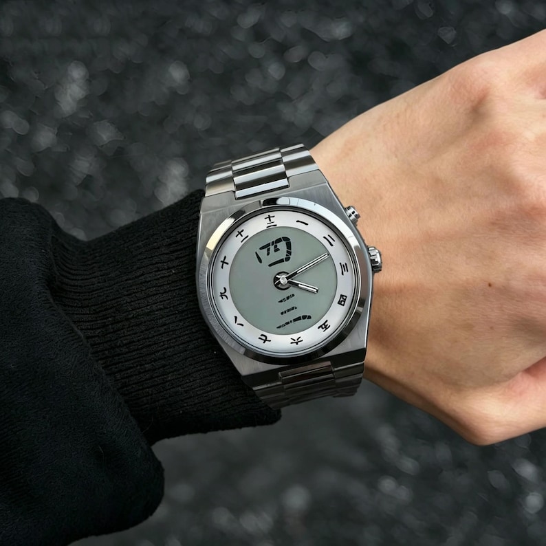 May include: A silver-toned stainless steel wristwatch featuring a light gray circular dial with black Japanese kanji numerals as hour markers. The watch includes a metallic link bracelet, silver hands, and a prominent kanji character at the center.