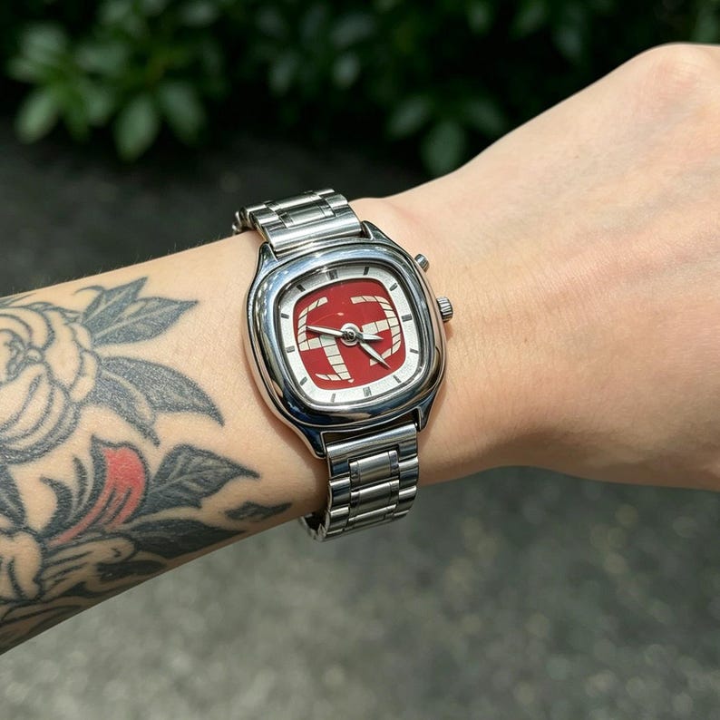 Puede incluir: Un reloj de pulsera plateado con una esfera cuadrada y un dise&ntilde;o rojo y blanco. El reloj tiene una correa plateada y un peque&ntilde;o dial en el lateral. La esfera del reloj presenta un dise&ntilde;o rojo con detalles en blanco.