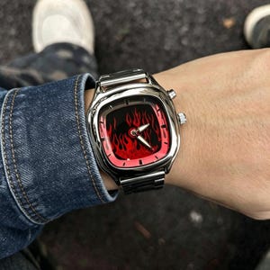 May include: A silver-toned wristwatch with a square-shaped case and a metallic link band. The watch face features a vibrant red and black flame pattern with silver-colored hour and minute hands, and two small circular buttons on the side of the case.