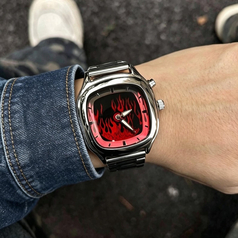 May include: A silver-toned wristwatch with a square-shaped case and a metallic link band. The watch face features a vibrant red and black flame pattern with silver-colored hour and minute hands, and two small circular buttons on the side of the case.