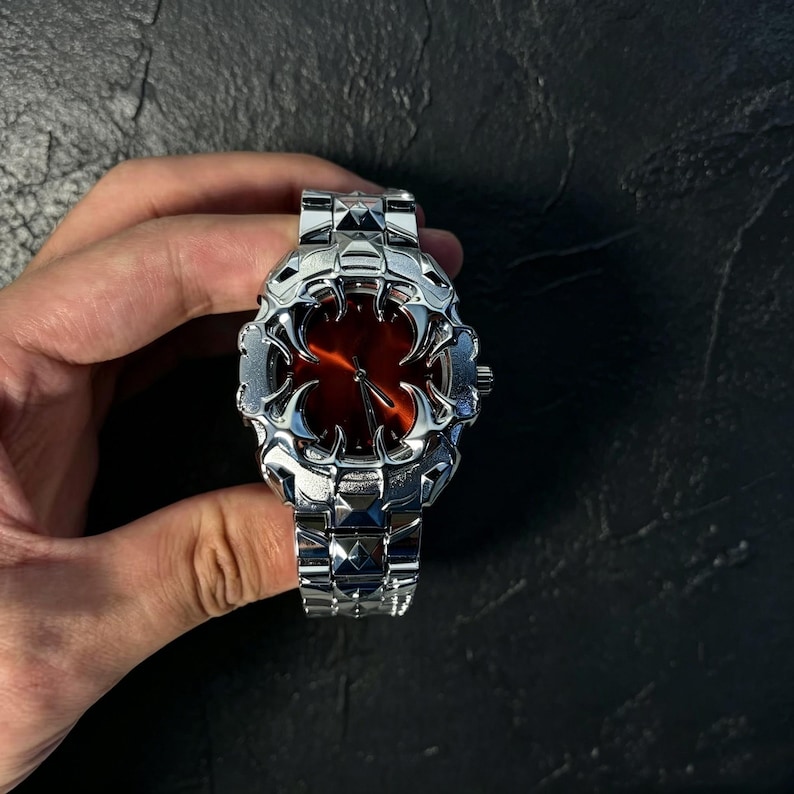 May include: A silver-toned wristwatch with a unique, spiky bezel and band. The watch face is a deep red color. The watch is held in a hand against a dark background. The watch band is made of interlocking metal links.