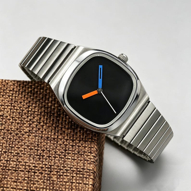 May include: A modern wristwatch with a silver-tone metal band and a black square watch face. The watch face features blue and orange accent markers and a white second hand. The watch is displayed on a brown textured surface.